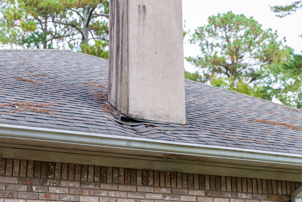 A brick chimney with water stains sits on a shingled roof that is sagging beneath it, with scattered pine needles and a gutter along the edge.