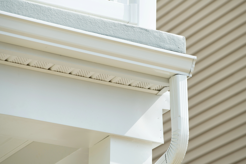 Close-up view of a white house exterior corner showing a roof gutter, soffit vents, and a downspout against beige siding.