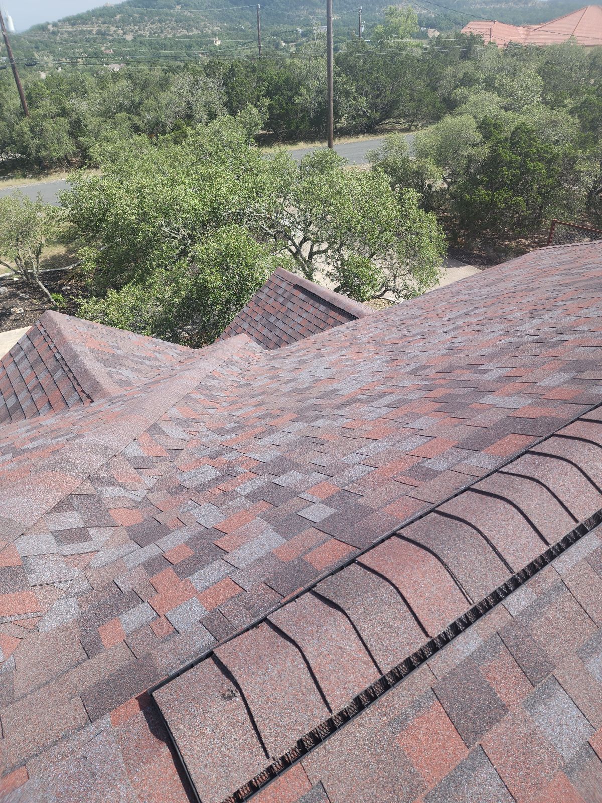 View of a house roof with reddish and gray asphalt shingles, surrounded by trees and overlooking a road and hills in the background.