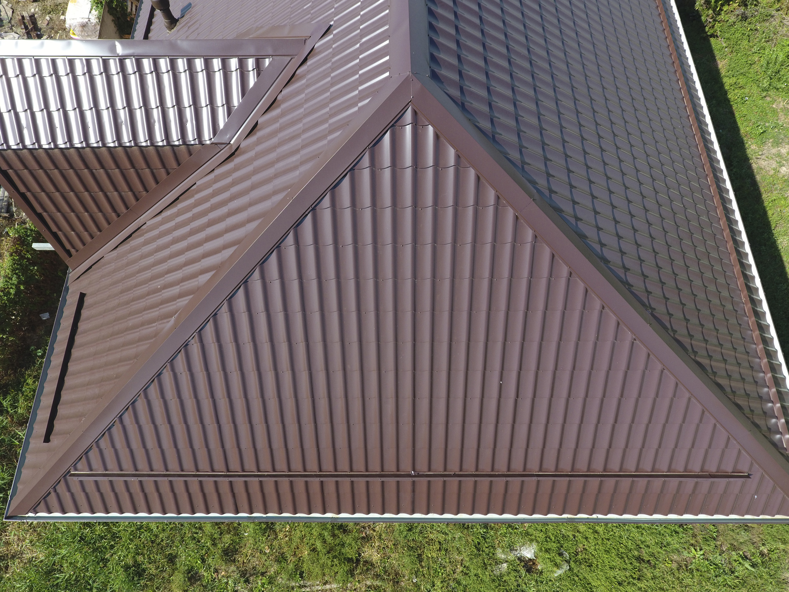Aerial view of a house roof with brown metal sheets and distinct ridges, surrounded by grass.