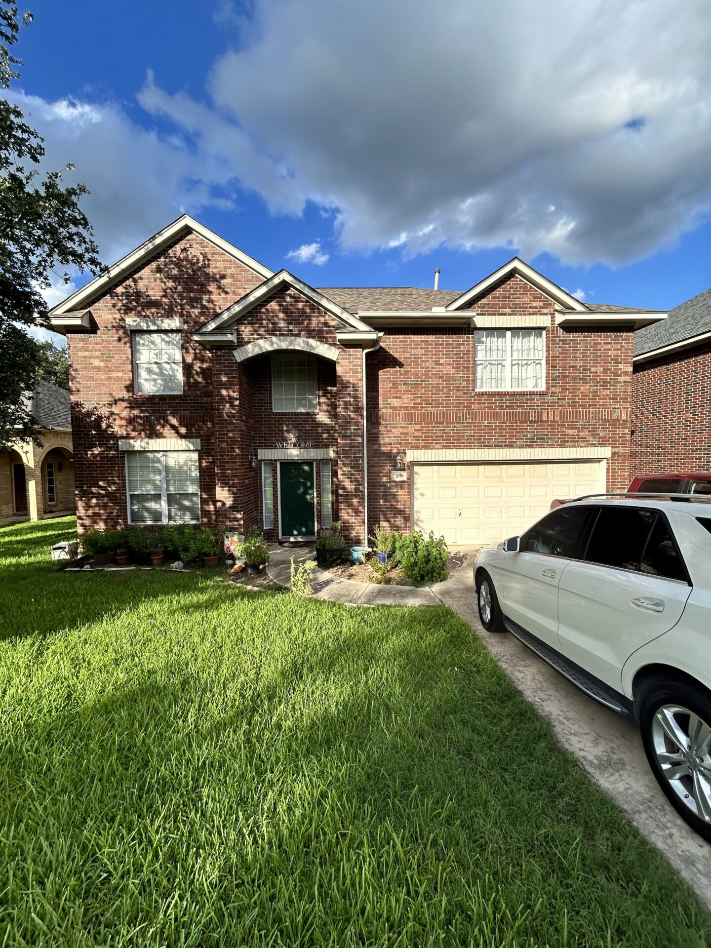 Two-story red brick house with attached two-car garage, white SUV parked in driveway, and green lawn in the foreground under a partly cloudy sky.