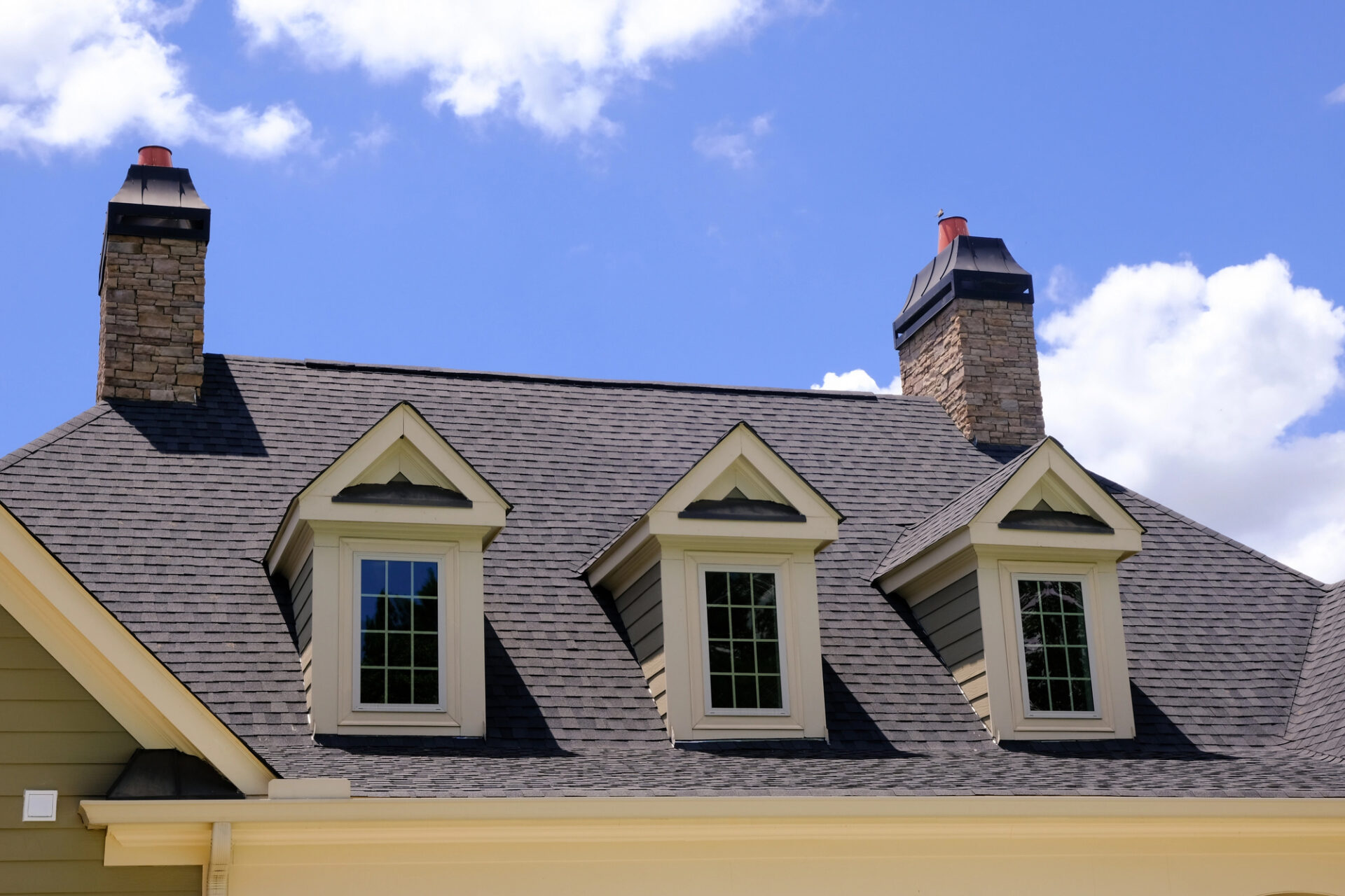 House roof with three dormer windows, two stone chimneys, gray shingles, and a blue sky with clouds—a perfect example of sturdy construction ideal for storm damage roof repair.