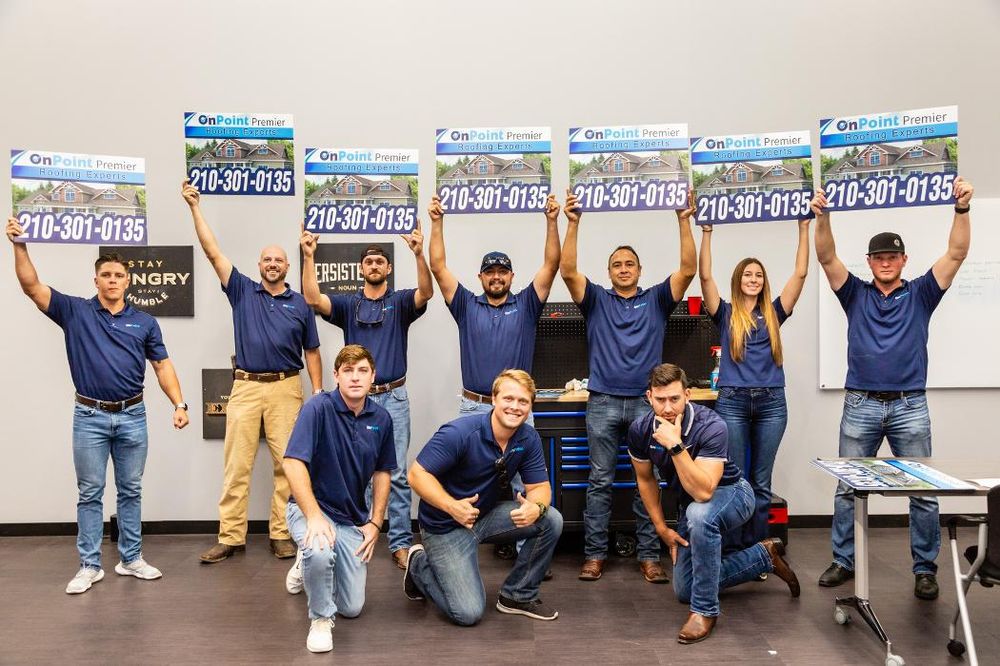 A group of nine people in blue shirts pose indoors, with five holding large OnPoint Premier Roofing signs displaying a phone number.